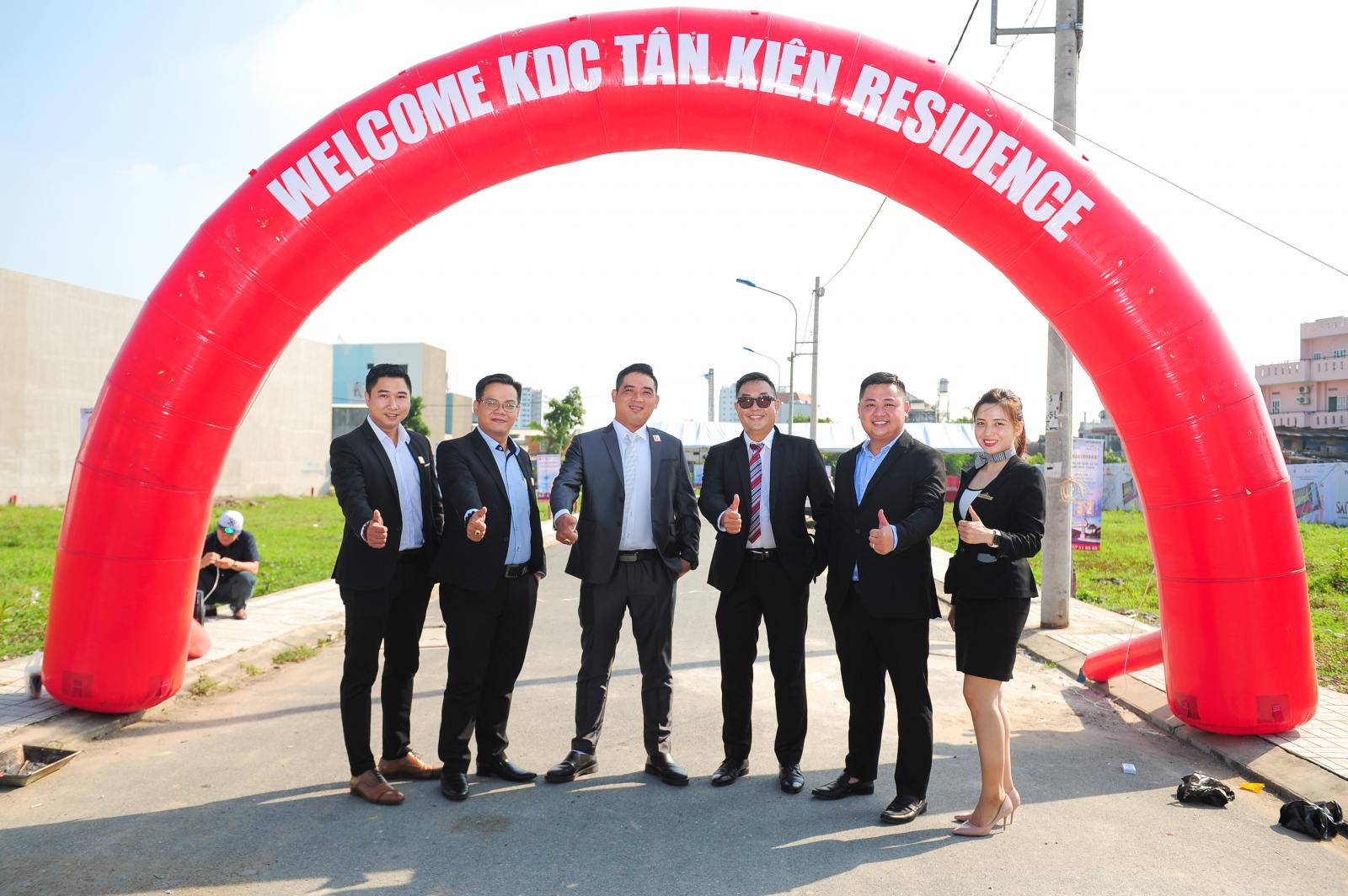5 người đàn ông và 1 phụ nữ mặc vest chỉnh tề chụp ảnh tại cổng chào màu đỏ có dòng chữ màu trắng Welcome KDC Tân Kiên Residence