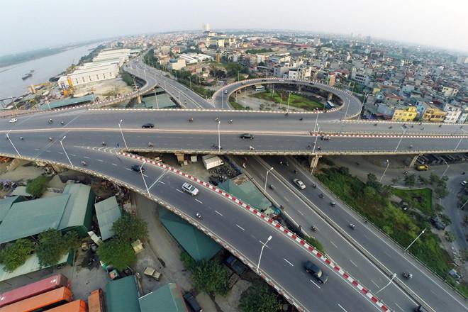 Nút giao cầu Vĩnh Tuy nhìn từ trên cao với nhiều làn đường và xe cộ đang di chuyển, xung quanh là nhiều công trình nhà ở và sông Hồng