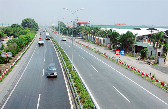 Tuyến cao tốc có nhiều ô tô đang chạy, ở giữa có hàng đèn đường, hai bên đường có nhà và cây cối