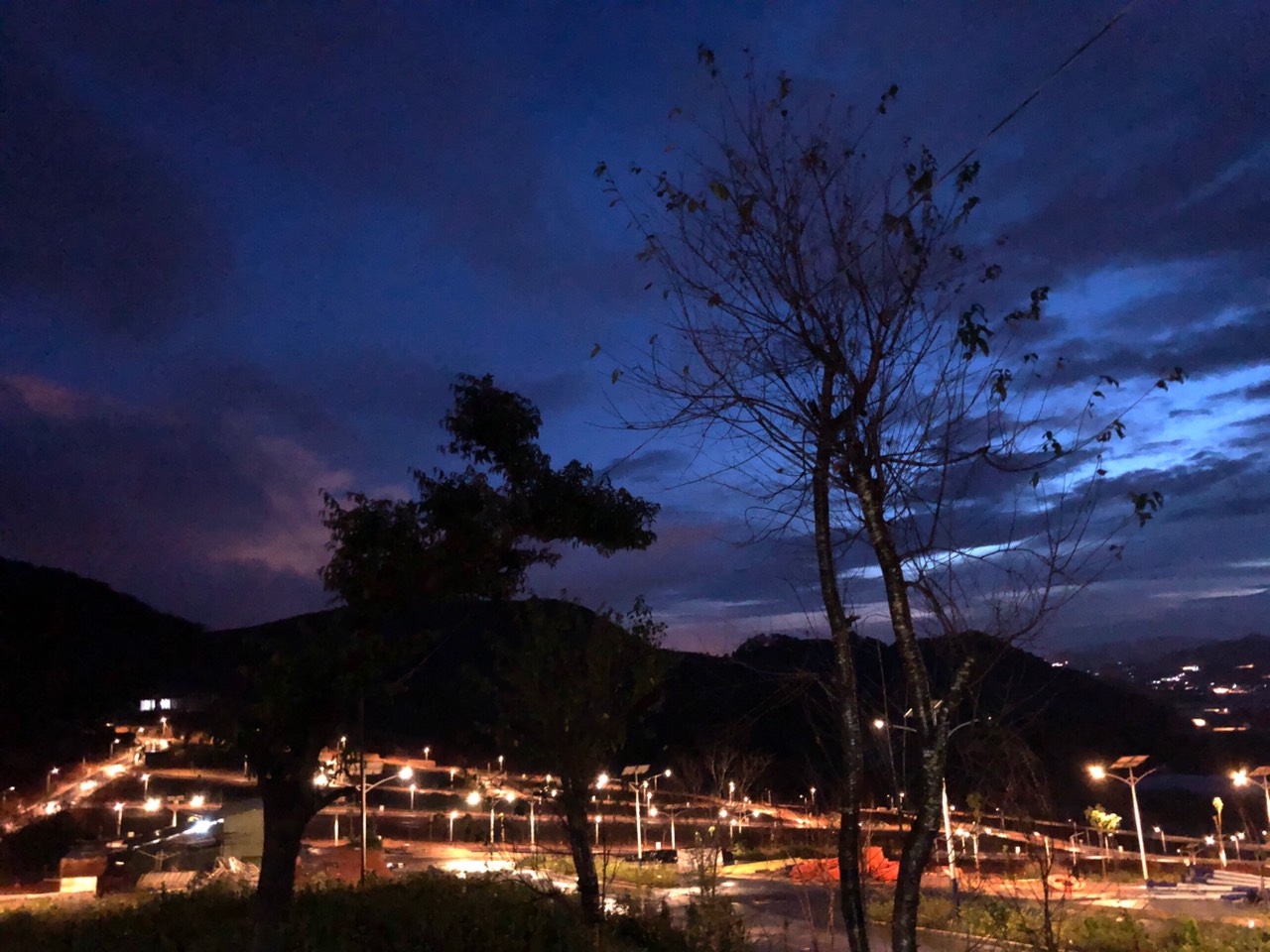 Cảnh bầu trời về đêm với nhiều đám mây, dưới mặt đất có nhiều cây xanh và bóng đèn đang thắp sáng